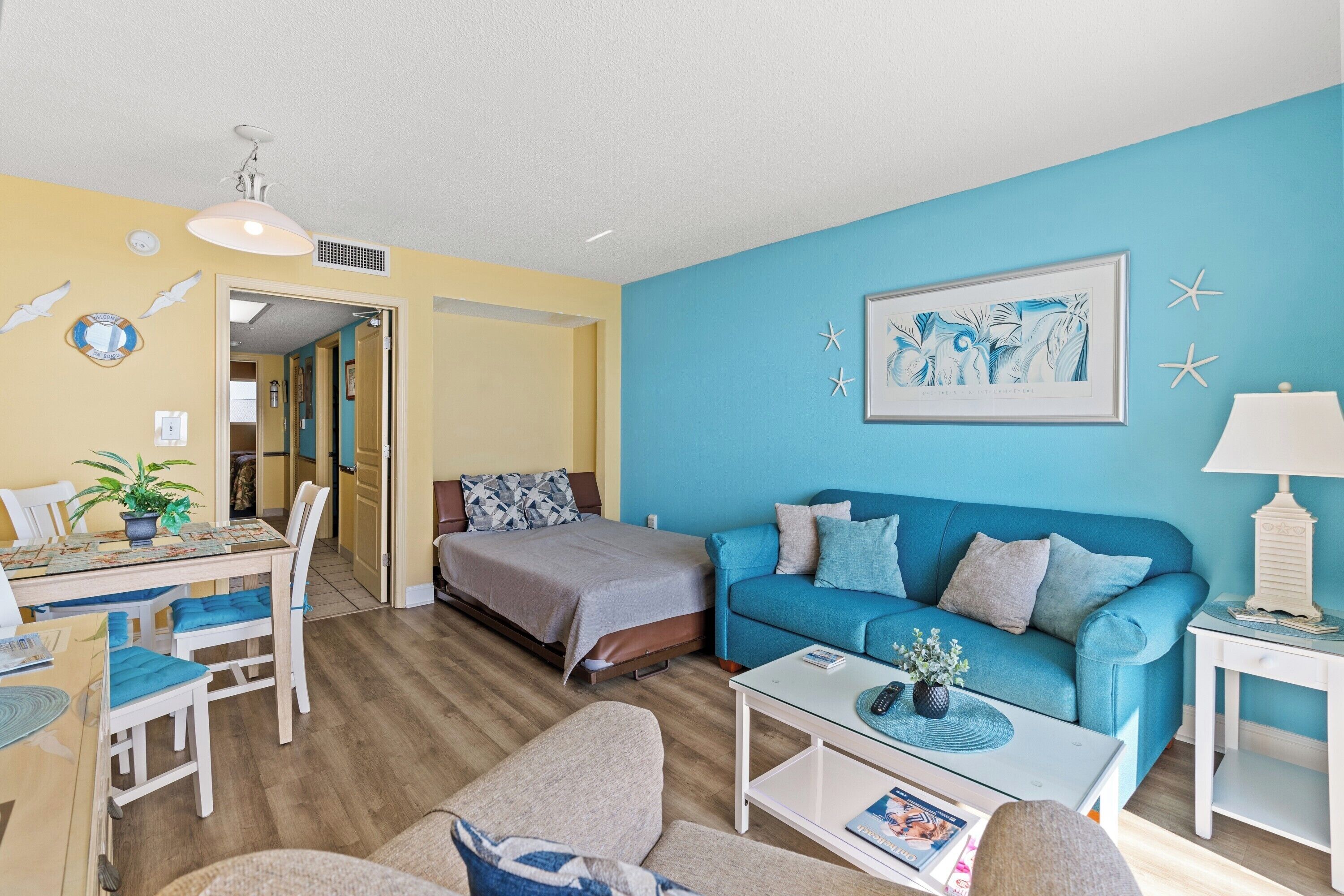A room with a bed, a blue sofa, a coffee table, and a dining area with chairs and a table.