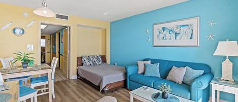 A room with a bed, a blue sofa, a coffee table, and a dining area with chairs and a table.