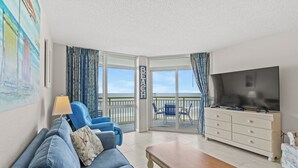 A living room with a sofa, coffee table, and a TV. There is a balcony with a view of the ocean.