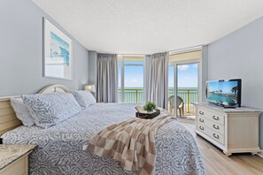 A hotel room with a bed, a TV, a desk with drawers, a window with curtains, and a view of the sea. - Bay Watch 805-3BR/2BA Oceanfront Condo in North Myrtle Beach (North Myrtle Beach)