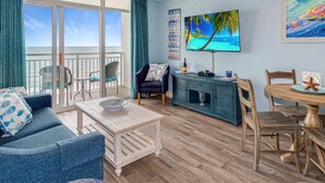 A living room with a sofa, a coffee table, a TV, a cabinet, and a dining table with chairs. There is a balcony with a view of the ocean. - Bay Watch 935-1BR/1BA Oceanfront Condo in North Myrtle Beach (North Myrtle Beach)