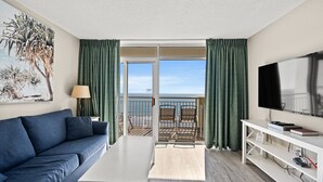 Ein Hotelzimmer mit Sofa, Fernseher, Tisch und einem Balkon mit Blick auf den Strand.