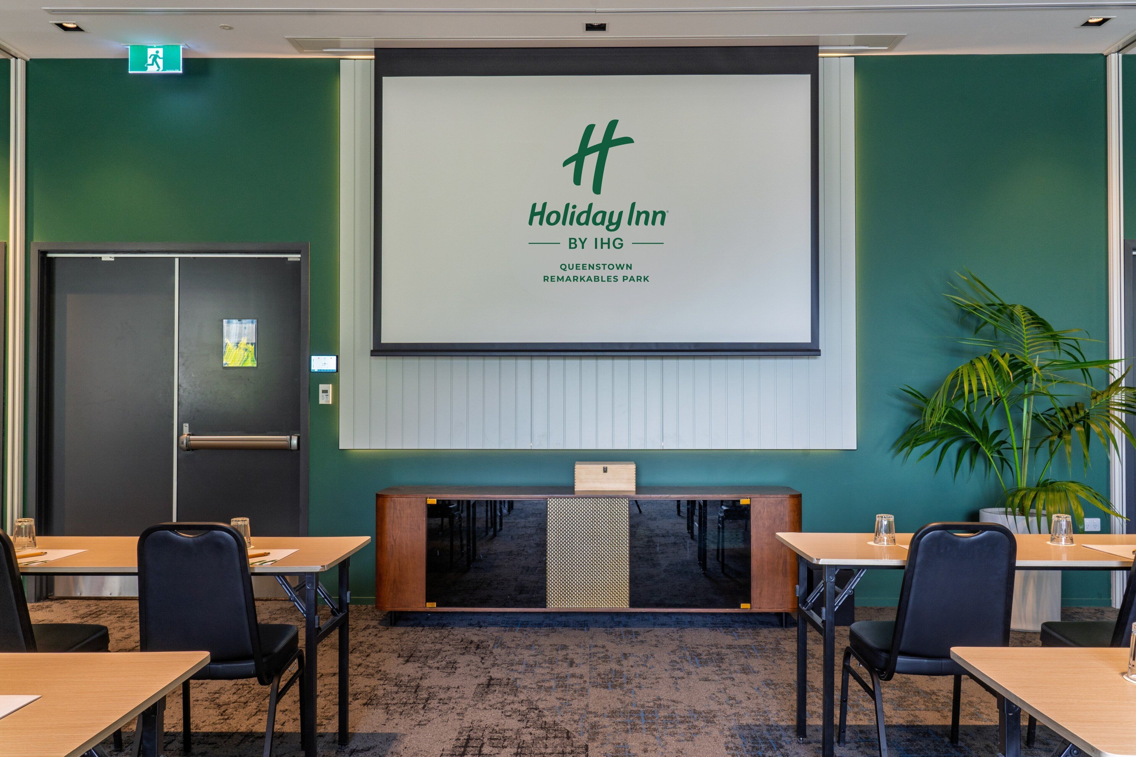 Ein Konferenzraum mit einer großen Leinwand, auf der das Holiday Inn Logo zu sehen ist, einem Empfangstresen und mehreren Tischen mit Stühlen.