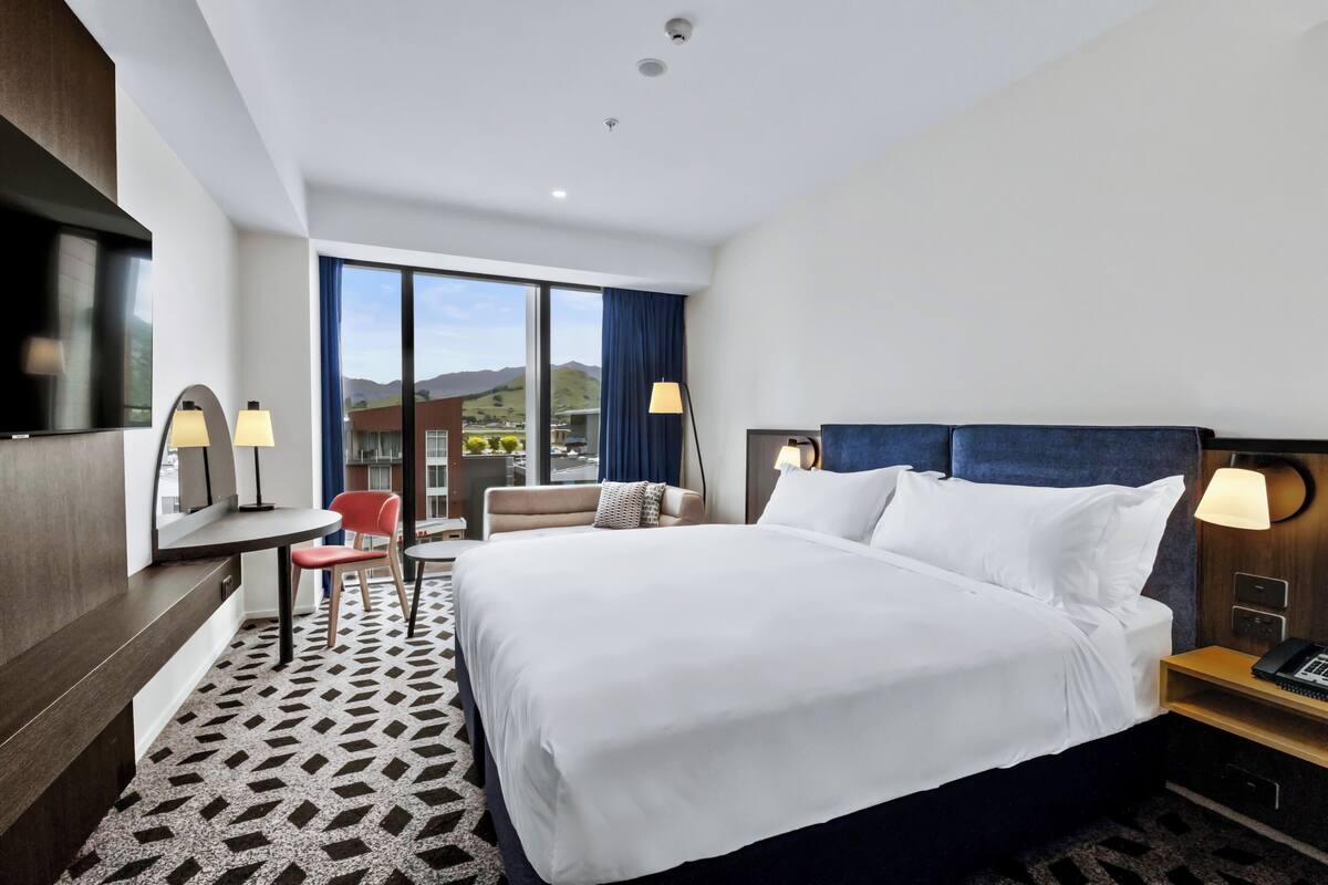 Une chambre d’hôtel avec un grand lit, un bureau avec une chaise, une télévision et une fenêtre donnant sur les montagnes.