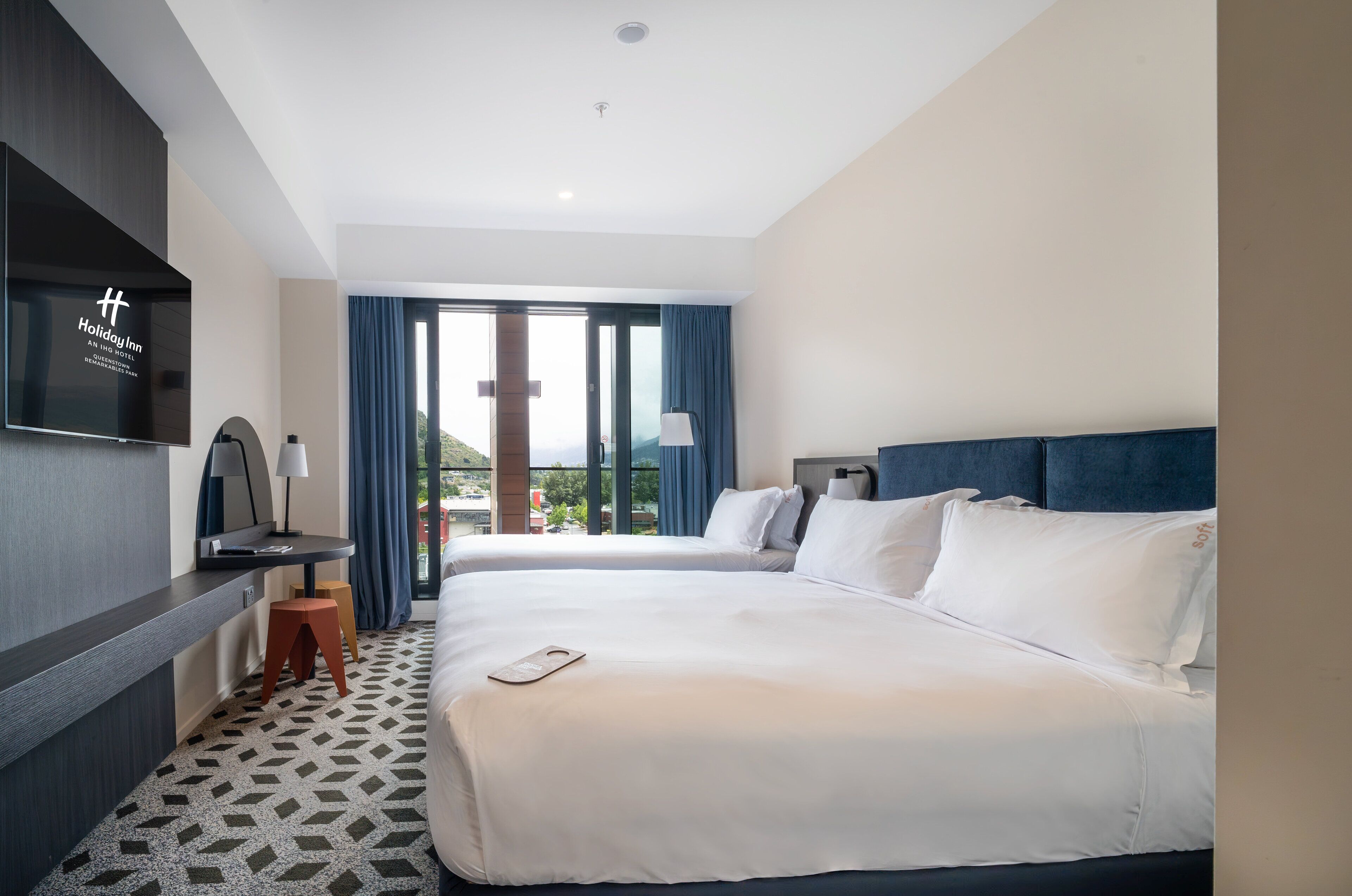 A hotel room with two beds, a flat-screen TV, a desk with a lamp, and a view of the outdoors.