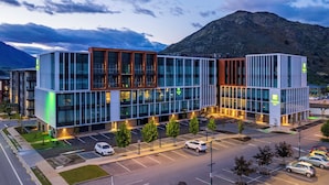A modern hotel with a glass facade, parking lot, and a mountainous backdrop. - Holiday Inn Queenstown Remarkables Park by IHG (Queenstown)