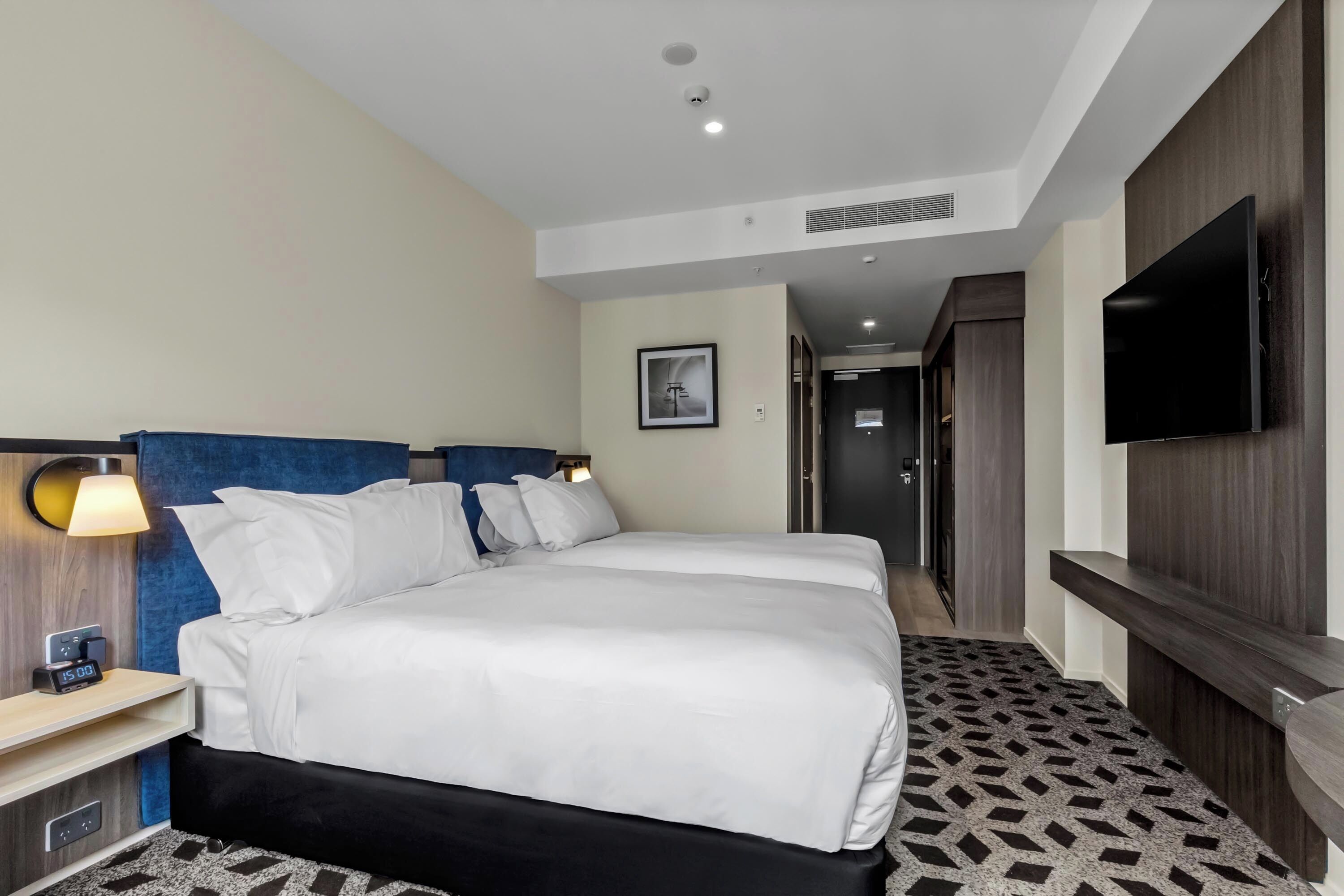 A hotel room with two beds, a TV, a desk with a lamp, and a patterned carpet.