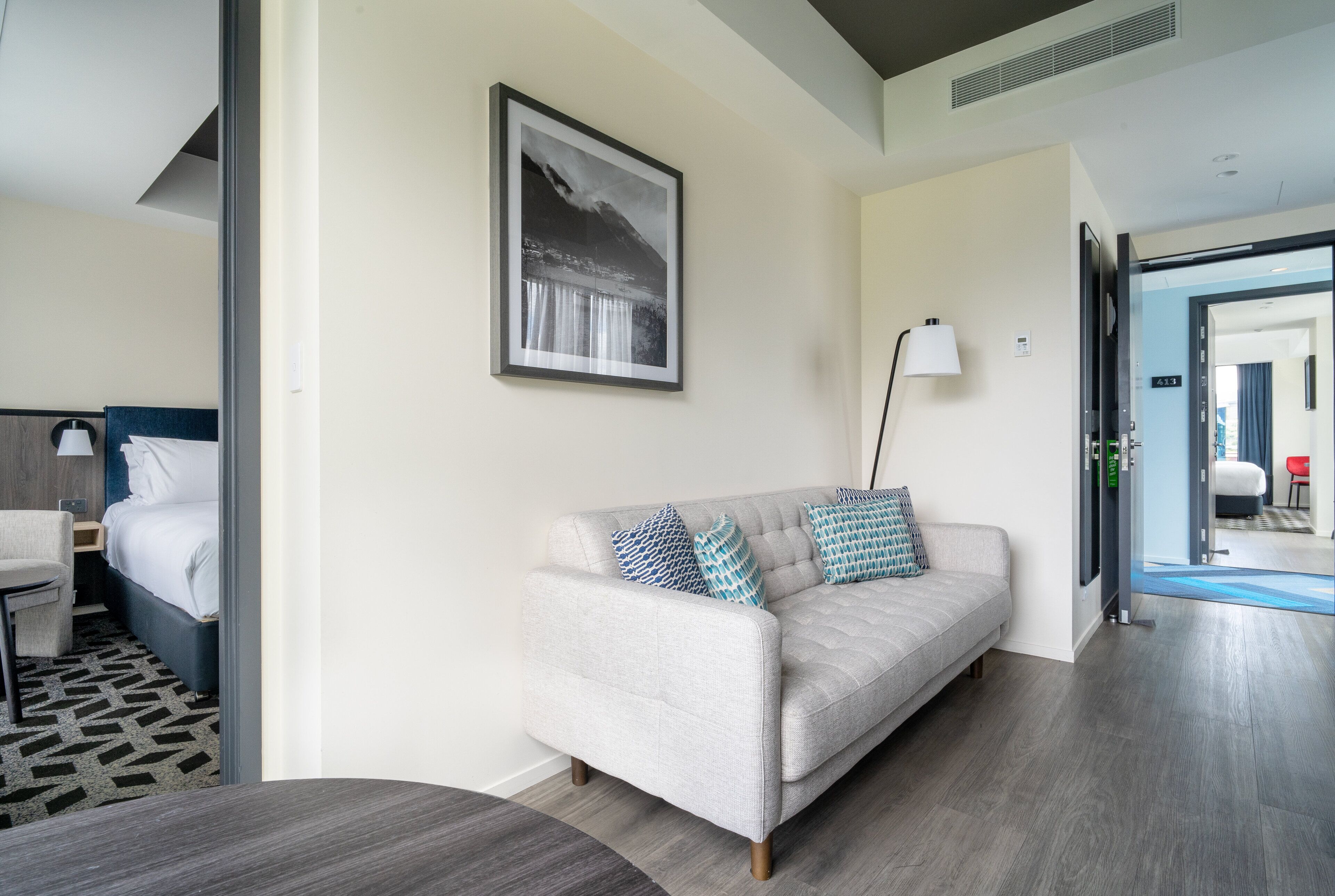 Una habitación de hotel moderna con un sofá gris, una cama, una mesita de noche, una lámpara y un cuadro enmarcado en la pared.