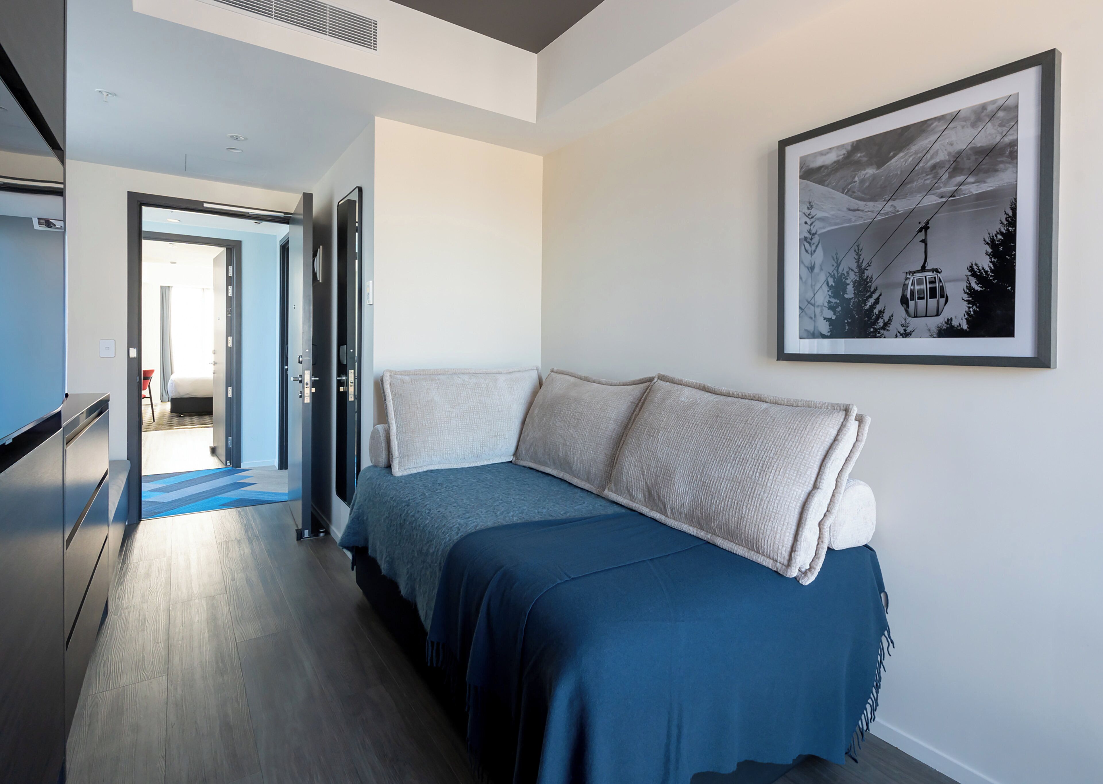 A modern hotel room with a bed, a framed picture on the wall, and a hallway leading to another room.