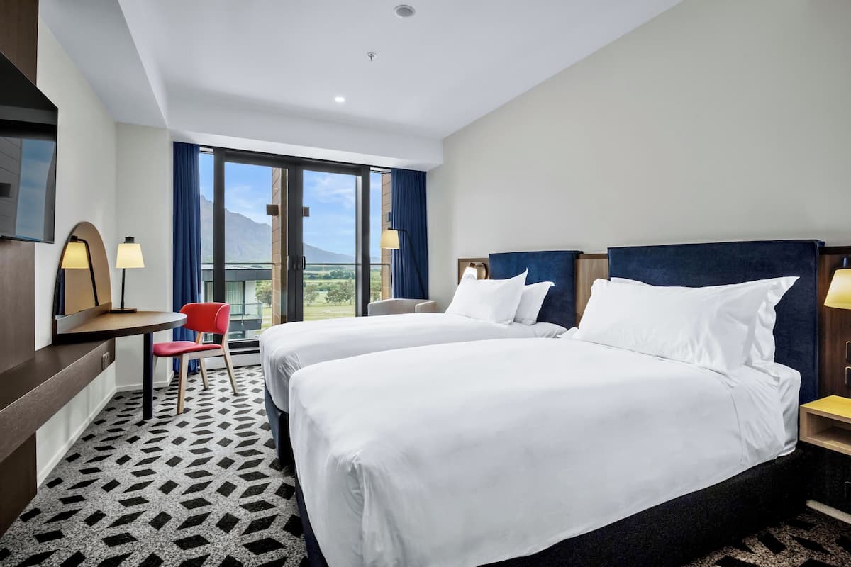 Une chambre d’hôtel avec deux lits, un bureau, une chaise et une grande fenêtre donnant sur un beau paysage.