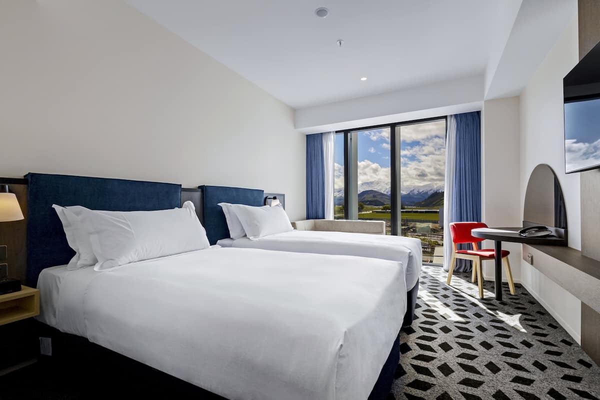Une chambre d’hôtel avec deux lits, un bureau, une chaise et une grande fenêtre donnant sur la montagne.