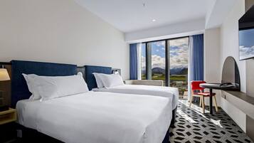 A hotel room with two beds, a desk, a chair, and a large window with a mountain view.
