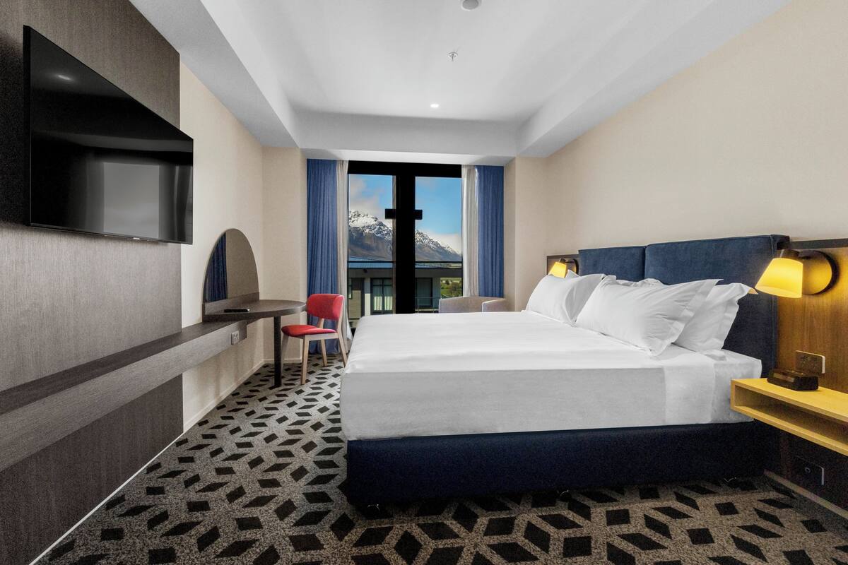 Une chambre d’hôtel avec un grand lit, un bureau, un téléviseur fixé au mur et une vue sur les montagnes par la fenêtre.