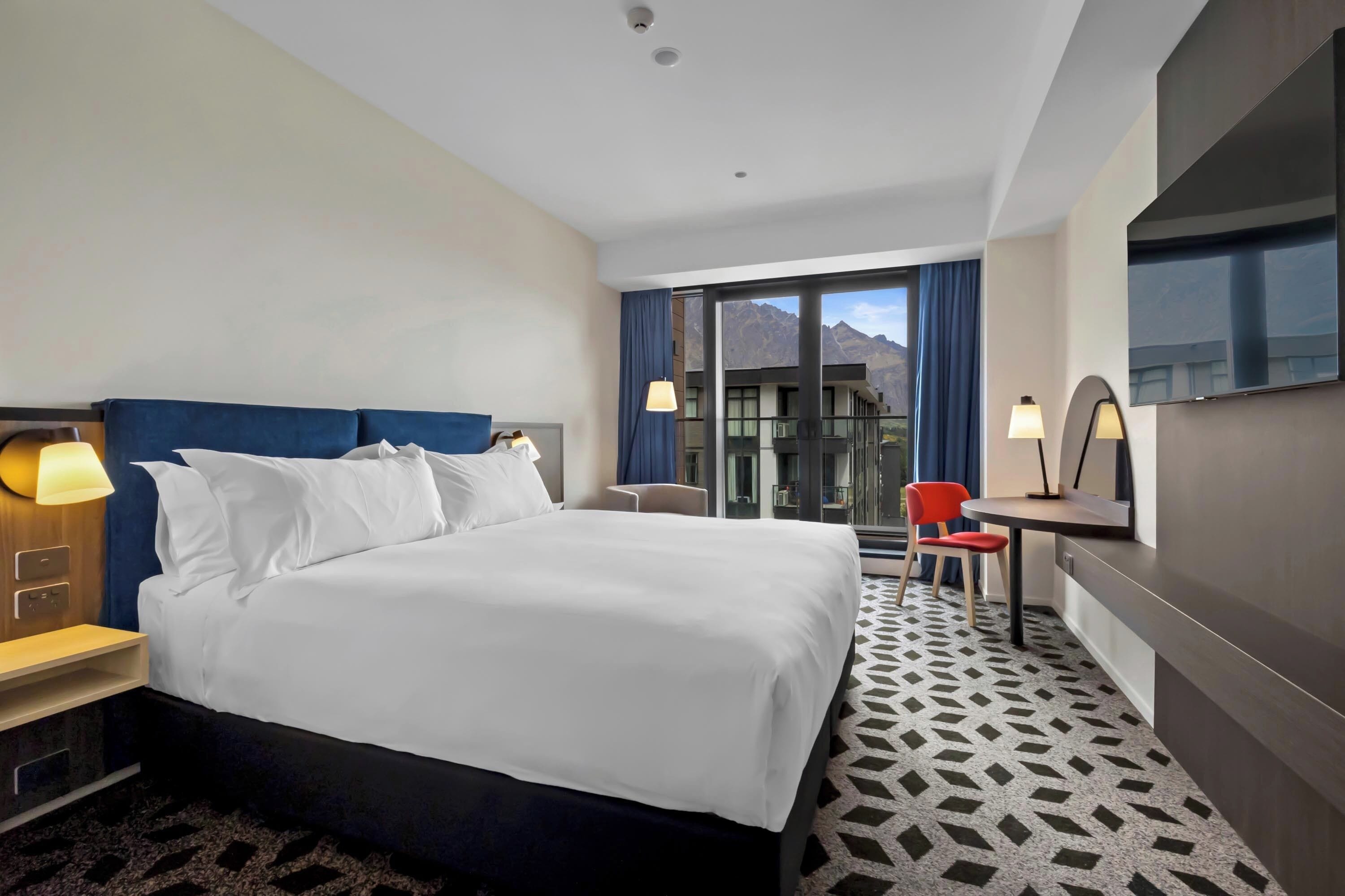 Habitación de hotel con una cama grande, un escritorio, una silla, un televisor y vistas a las montañas a través de la ventana.