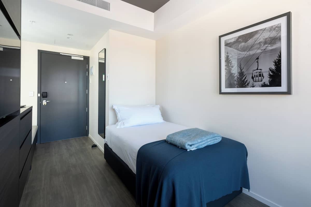 Une chambre d’hôtel avec un lit, une commode en bois sombre, une porte et un tableau représentant un téléphérique dans une montagne.