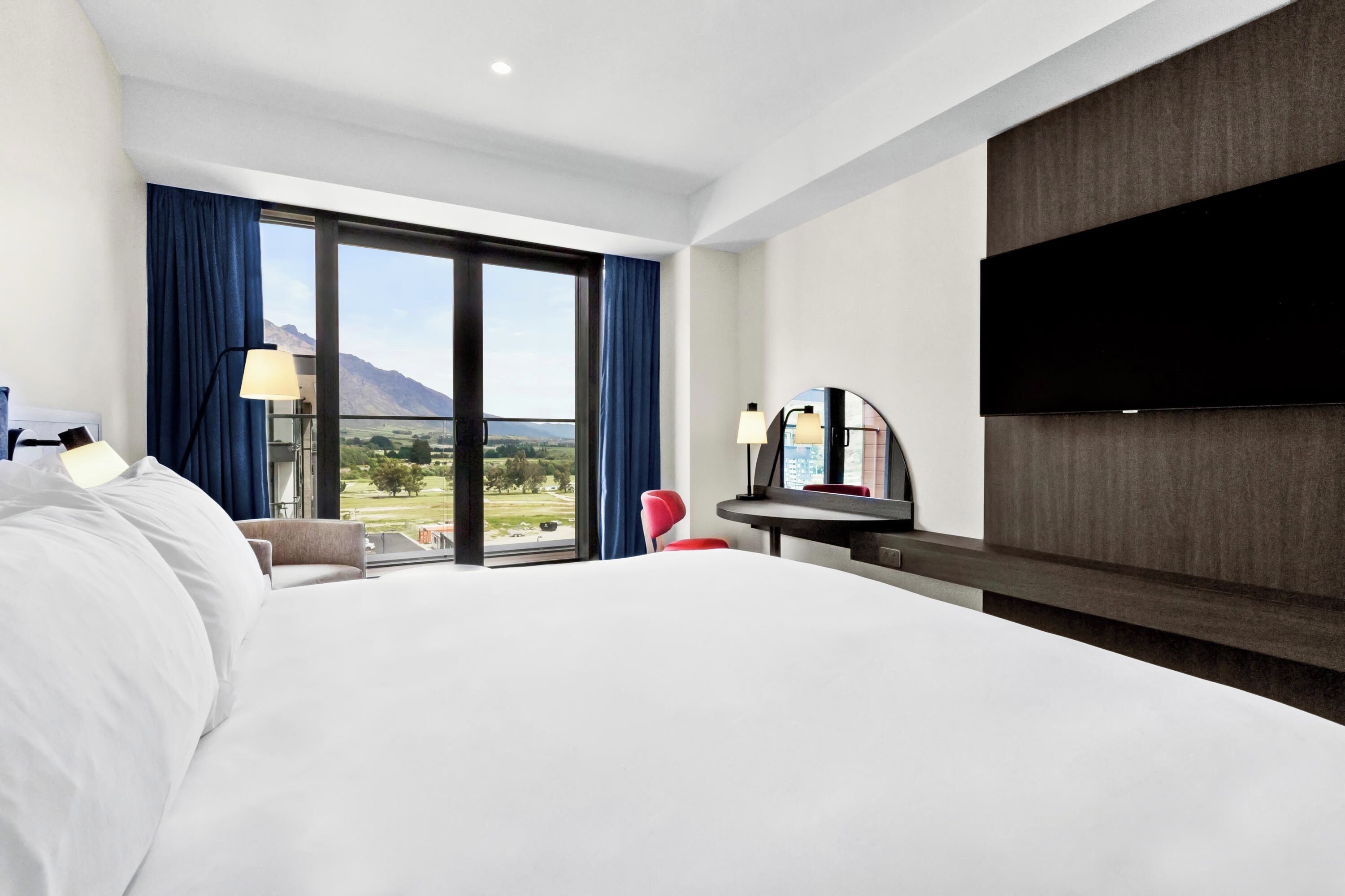 A hotel room with a large bed, a desk with a chair, a flat-screen TV, and a view of the outdoors.