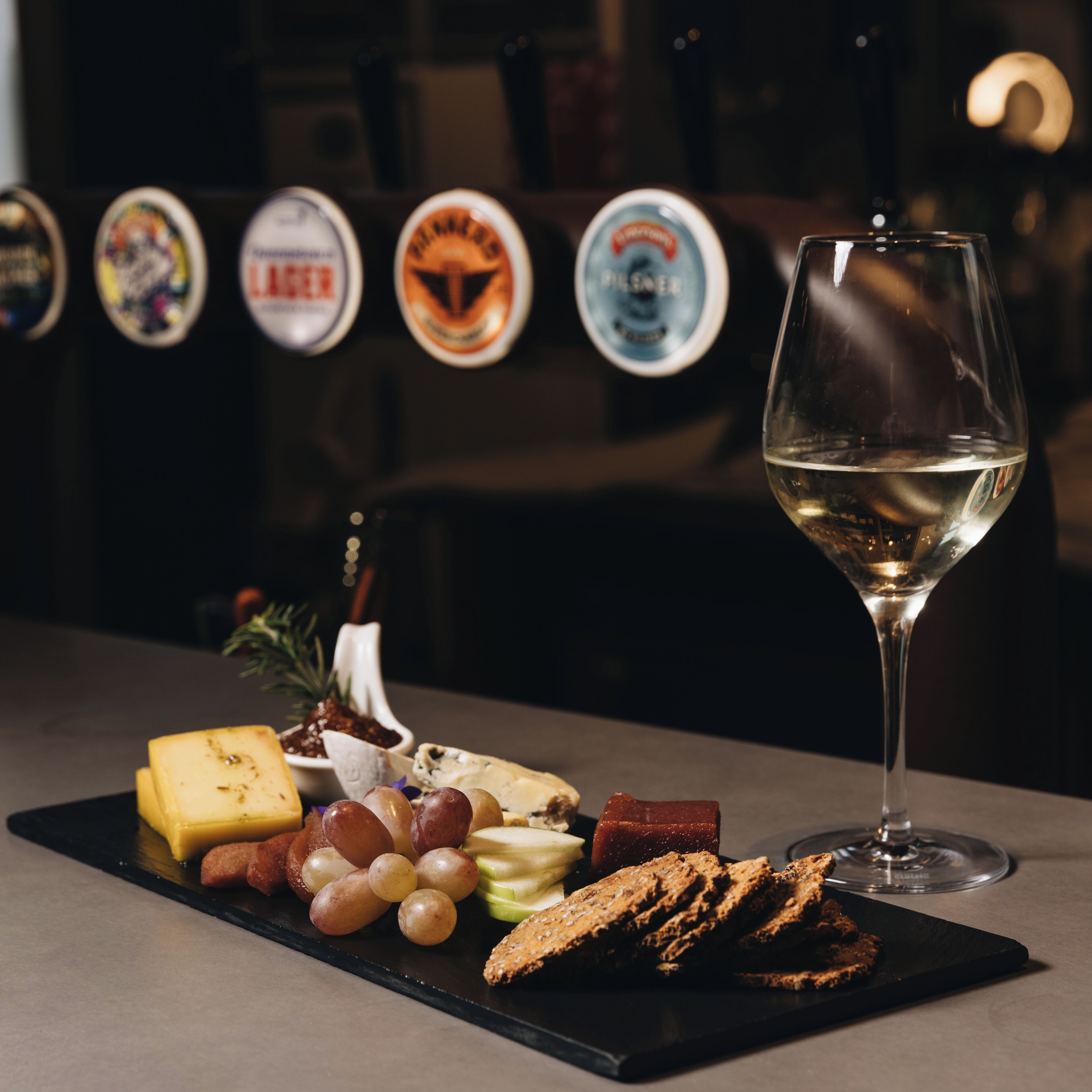 A selection of cheeses, fruits, and bread on a black tray, accompanied by a glass of white wine, set against a backdrop of beer taps.