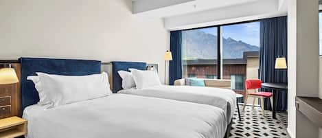 A hotel room with two beds, a desk, a chair, and a large window with a mountain view.