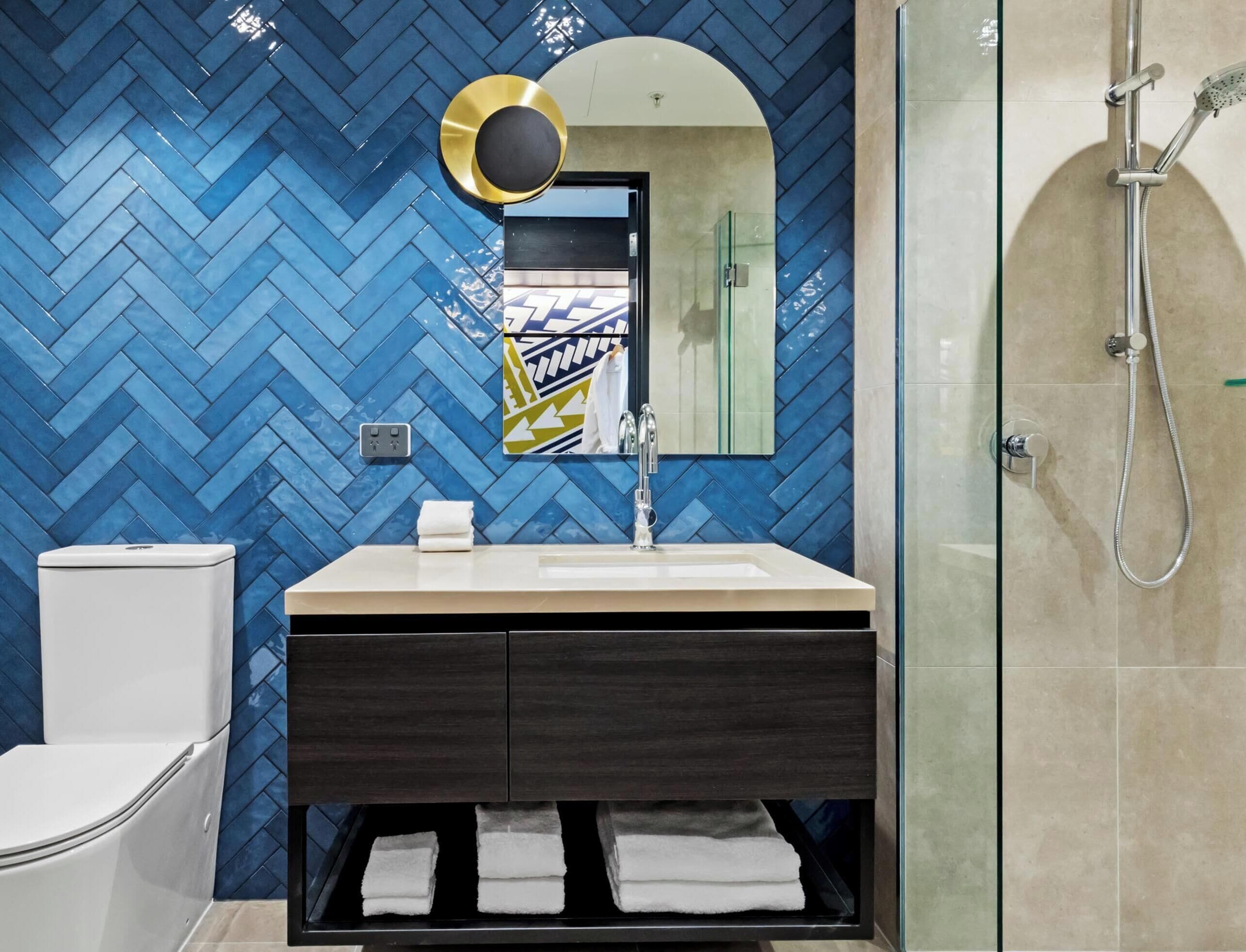 A bathroom with a herringbone tile wall, a modern sink with a mirror, a glass shower enclosure, and a toilet.