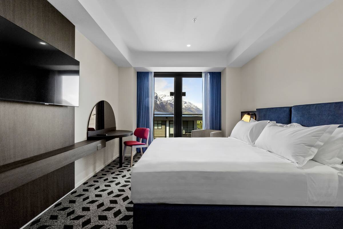 Une chambre d’hôtel avec un grand lit, un bureau, une chaise, un téléviseur fixé au mur et une vue sur les montagnes par la fenêtre.