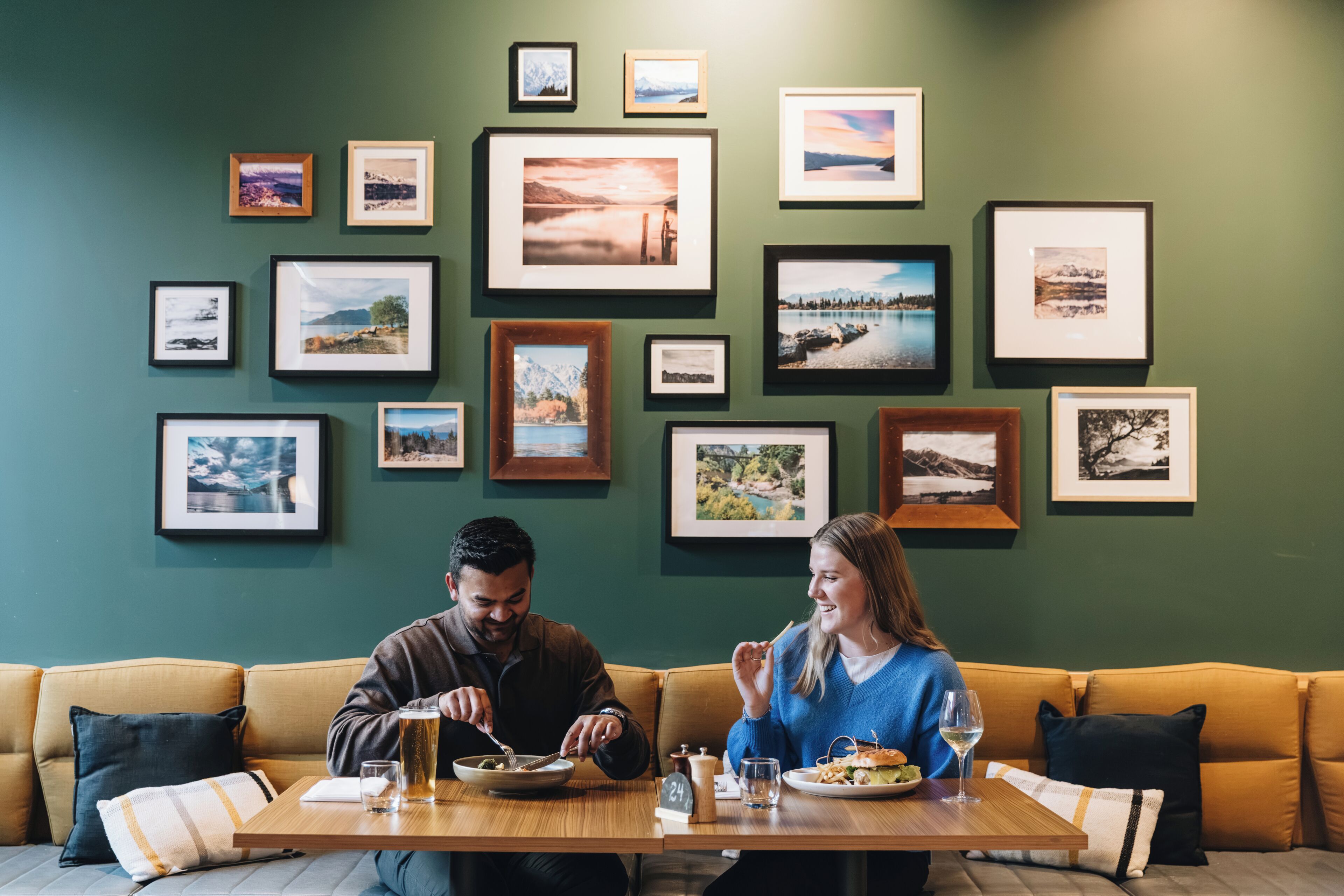Un comedor con una mesa de madera, dos personas sentadas y una pared decorada con fotografías enmarcadas de paisajes.