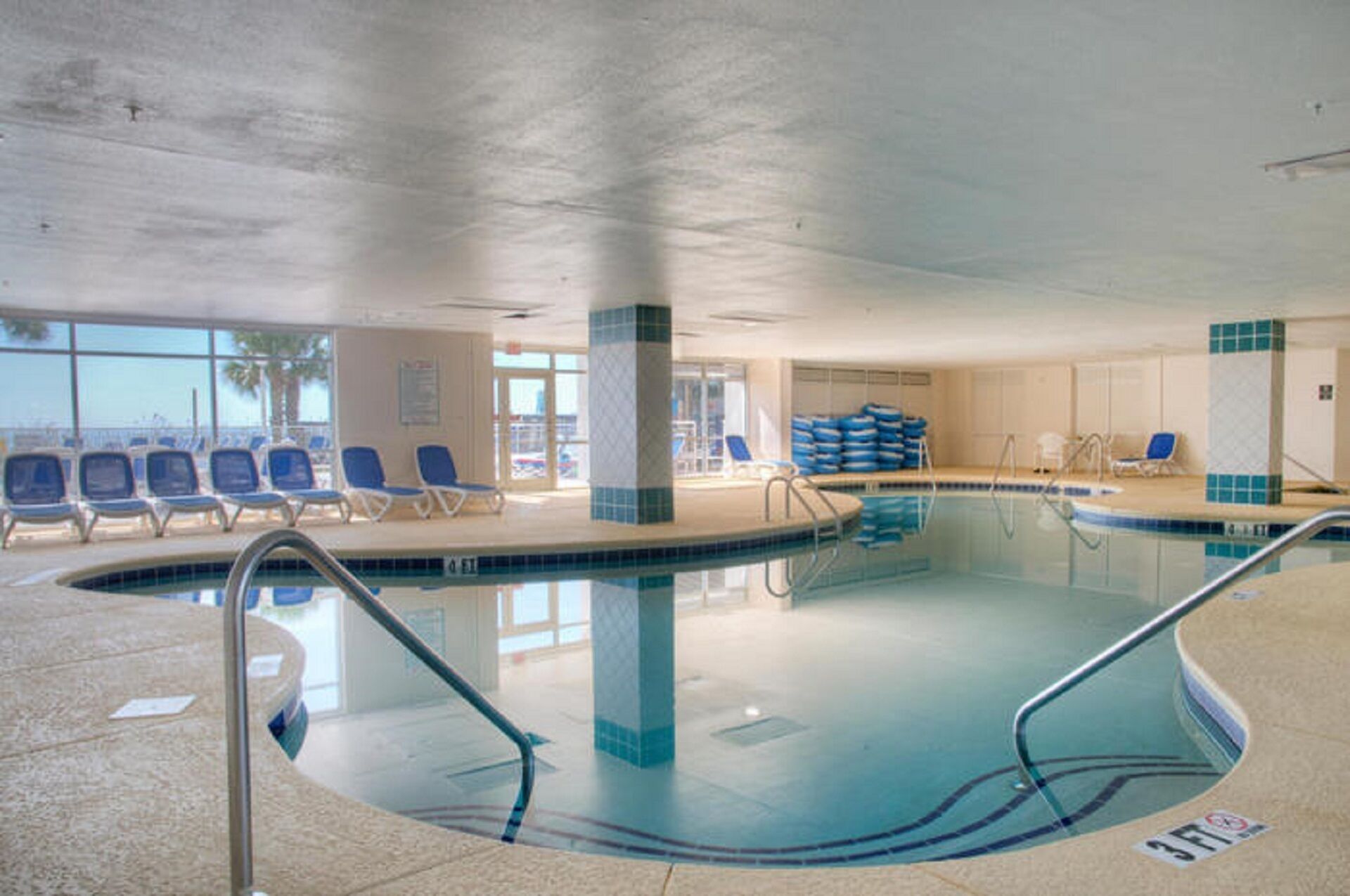 Une piscine intérieure à l’eau claire, avec des chaises bleues et un sol carrelé.