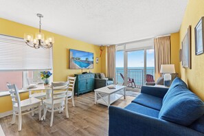 A living room with a sofa, dining table, chairs, a TV, and a balcony with a view.