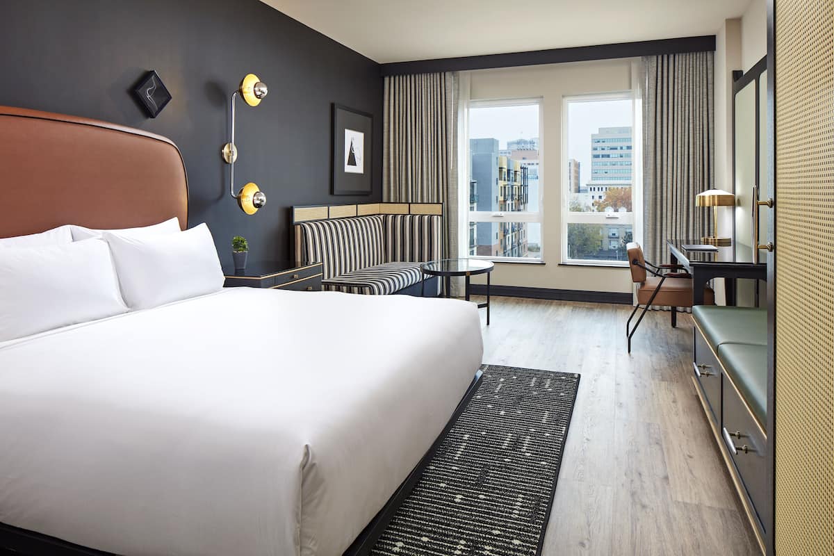 A modern hotel room with a large bed, a sofa, a small table, and a desk. There is a view of the city through the window.