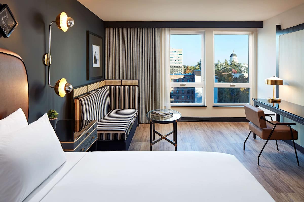 A modern hotel room with a large bed, a striped sofa, a small round table, and a desk with a chair. The room has a large window with a view of buildings and a dome structure.