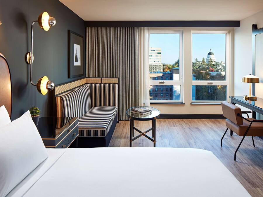 A modern hotel room with a large bed, a striped sofa, a small round table, and a desk with a chair. The room has a large window with a view of buildings and a dome structure.