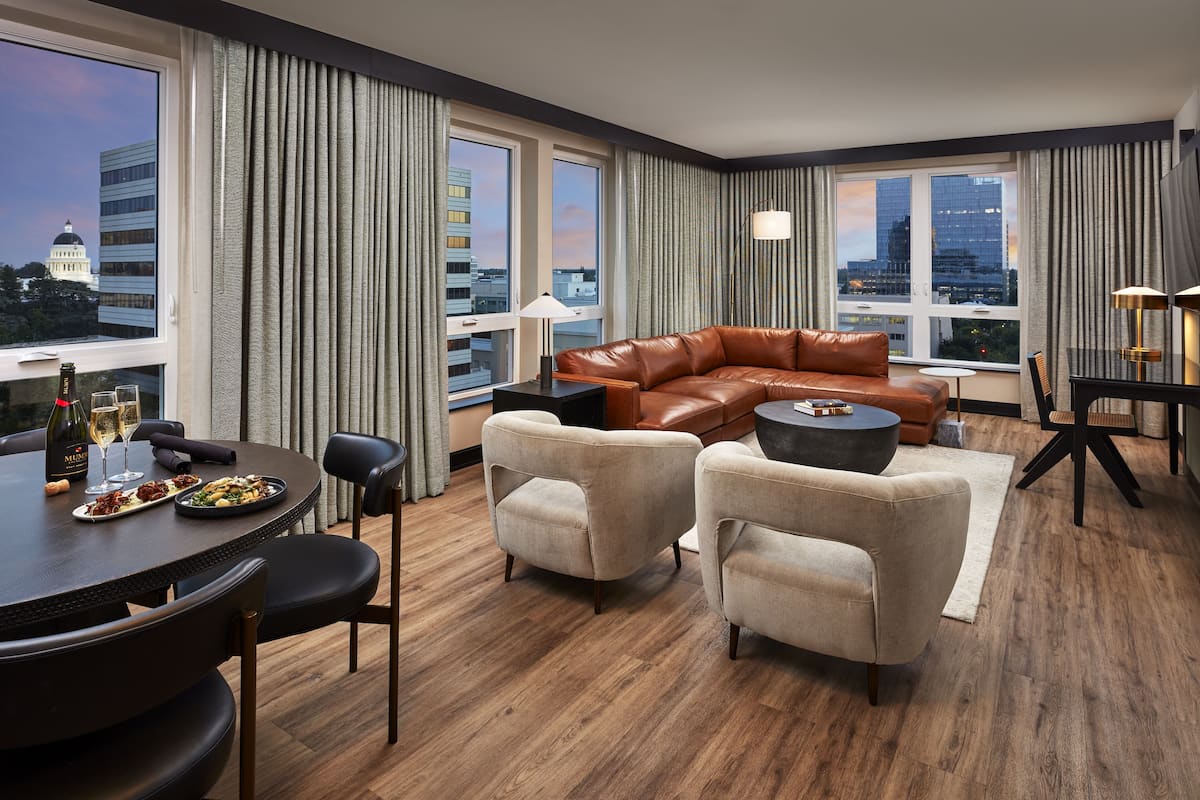 A modern hotel room with a leather sofa, two armchairs, a dining table with chairs, and large windows offering city views.