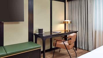 A hotel room with a desk, a chair, a green sofa, a TV, a lamp, and a window with curtains.