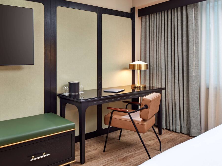 A hotel room with a desk, a chair, a green sofa, a TV, a lamp, and a window with curtains.