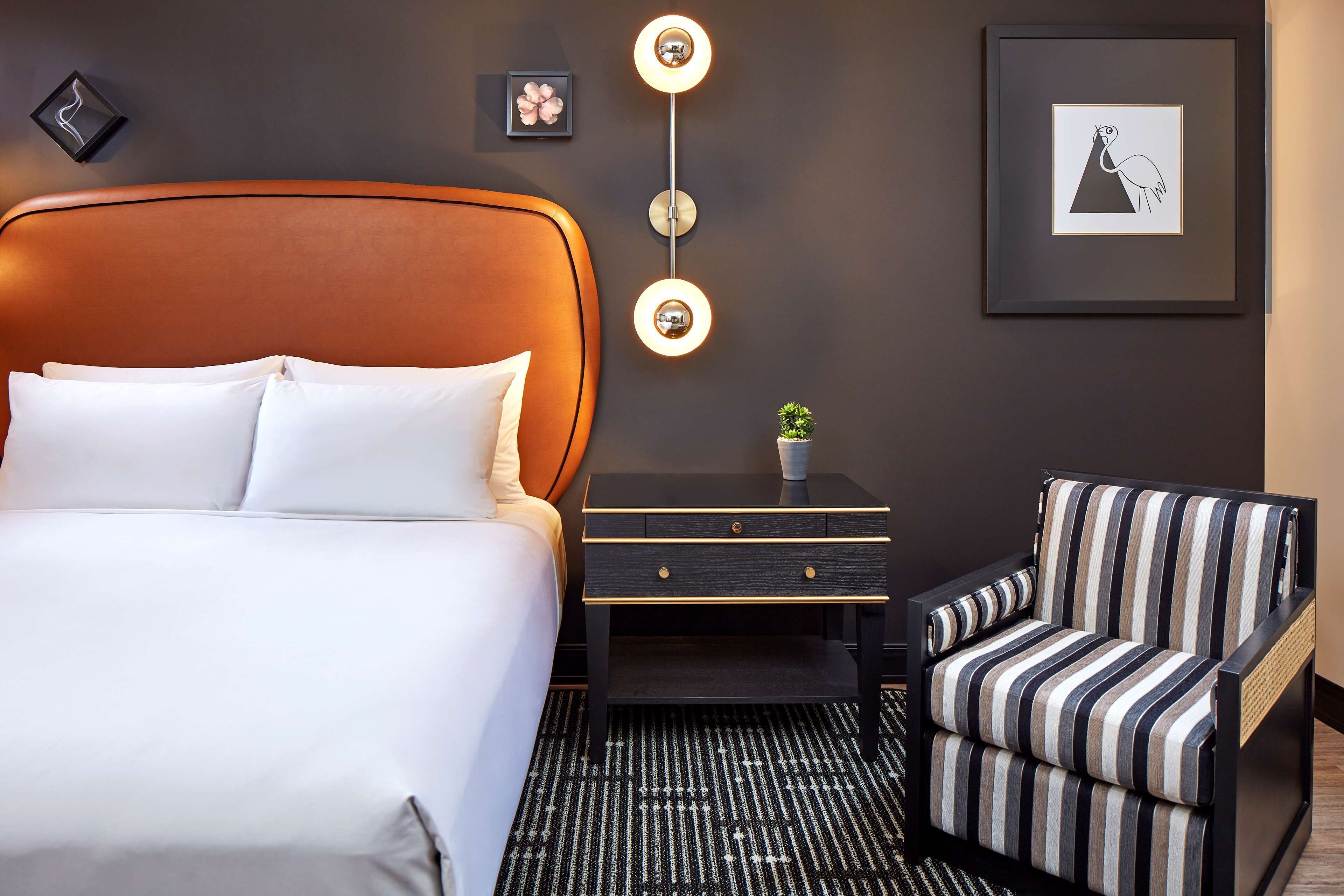 A modern hotel room with a large bed, a striped armchair, a nightstand, and a wall-mounted light fixture.