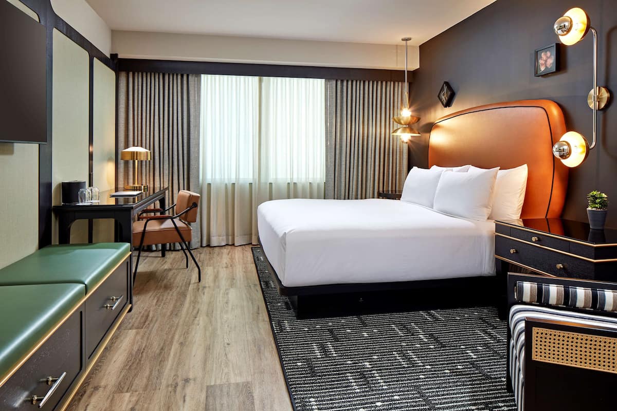A hotel room with a large bed, a desk with a lamp, a chair, and a small table.
