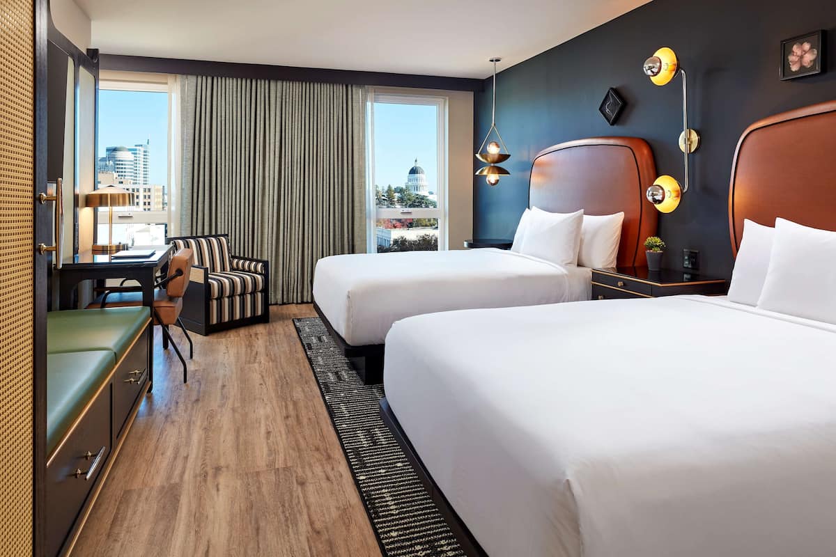 A hotel room with two beds, a desk, a chair, and a view of a cityscape.