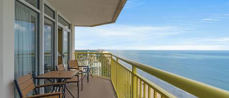 A balcony with tables and chairs, overlooking a beach and the ocean.