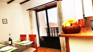 Un coin repas avec une table en verre, des chaises rouges, un banc en bois, un panier de fruits et une vue sur l’extérieur, avec un balcon visible à travers une fenêtre.