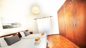 Une chambre d’hôtel avec deux lits, une armoire en bois, un rideau blanc et un luminaire suspendu.