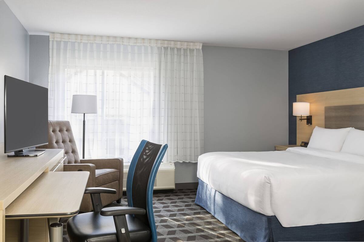 Ein Hotelzimmer mit einem Bett, einem Schreibtisch mit Computer, einem Sessel, einer Lampe und einem Fernseher.
