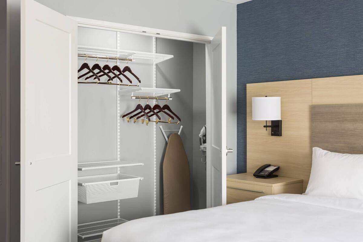 Ein Hotelzimmer mit einem ordentlich eingerichteten Kleiderschrank, einem Bett, einem Nachttisch mit Lampe und einem Telefon.