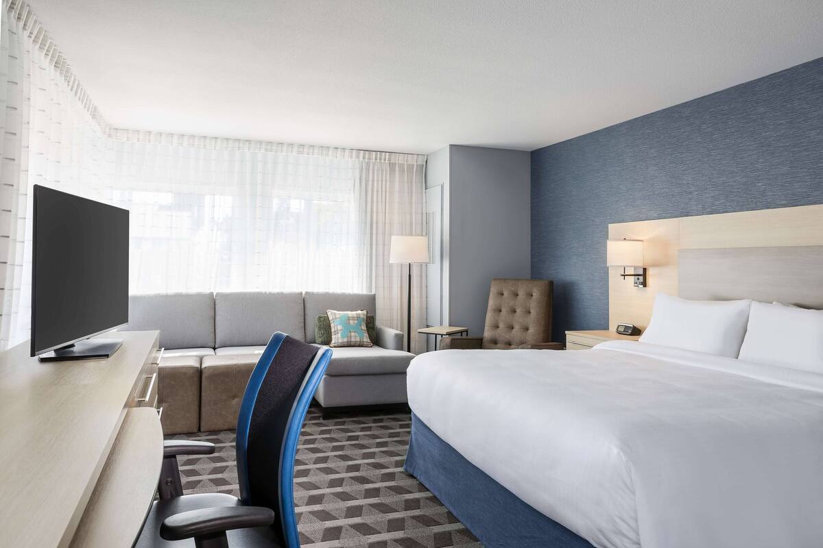 Ein Hotelzimmer mit einem Bett, einem Schreibtisch mit Fernseher, einer Couch, einem Sessel, einer Lampe und einem Nachttisch.