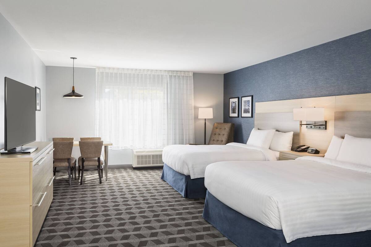 Ein Hotelzimmer mit zwei Betten, einem Schreibtisch mit Stühlen, einem Fernseher und einem Fenster mit Vorhängen.