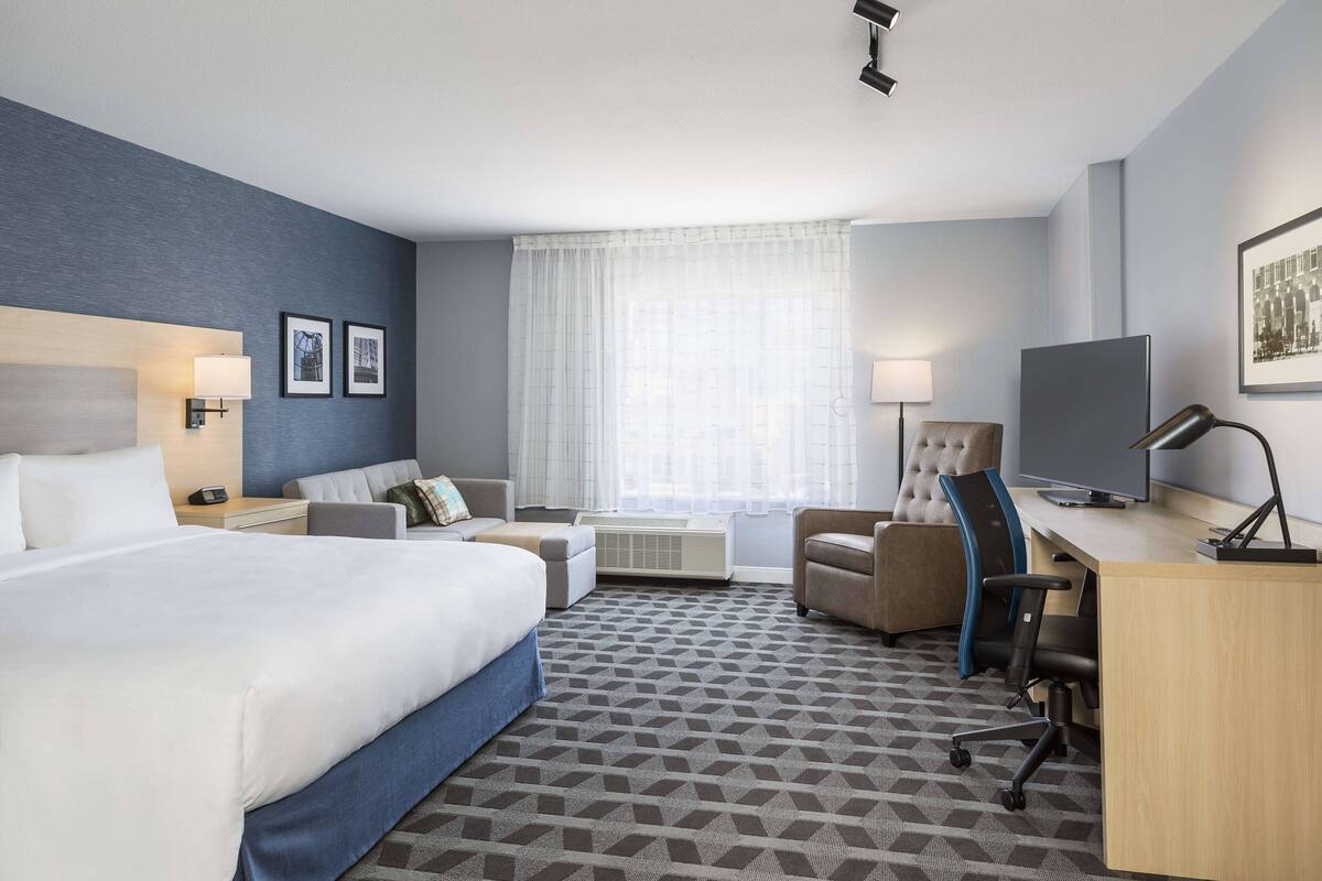 Ein Hotelzimmer mit einem großen Bett, einem Schreibtisch mit Computer, einem Sessel, einer Couch und einem Fernseher.