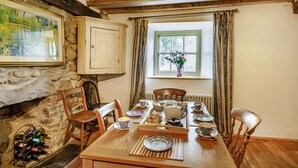 Ein Esszimmer mit Holztisch, gedeckt für vier Personen, umgeben von Stühlen, einem Kamin aus Stein und einem Fenster mit Vorhängen.
