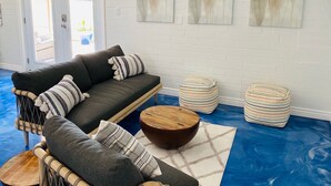 Un salon avec un canapé gris, des coussins à rayures, une table basse en bois et des poufs à rayures. Trois tableaux abstraits ornent le mur.