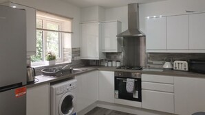 A modern kitchen with white cabinets, a stainless steel range hood, a washing machine, and a window with blinds.