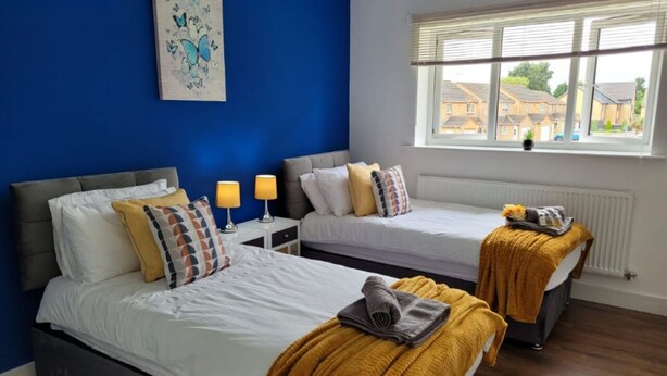 A hotel room with two beds, a window with a view of houses, and a blue wall with a butterfly painting.