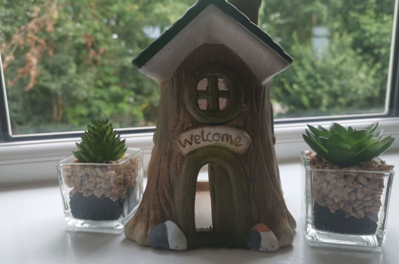 A wooden birdhouse with a 'welcome' sign, two small potted succulents, and a window with a view of trees.