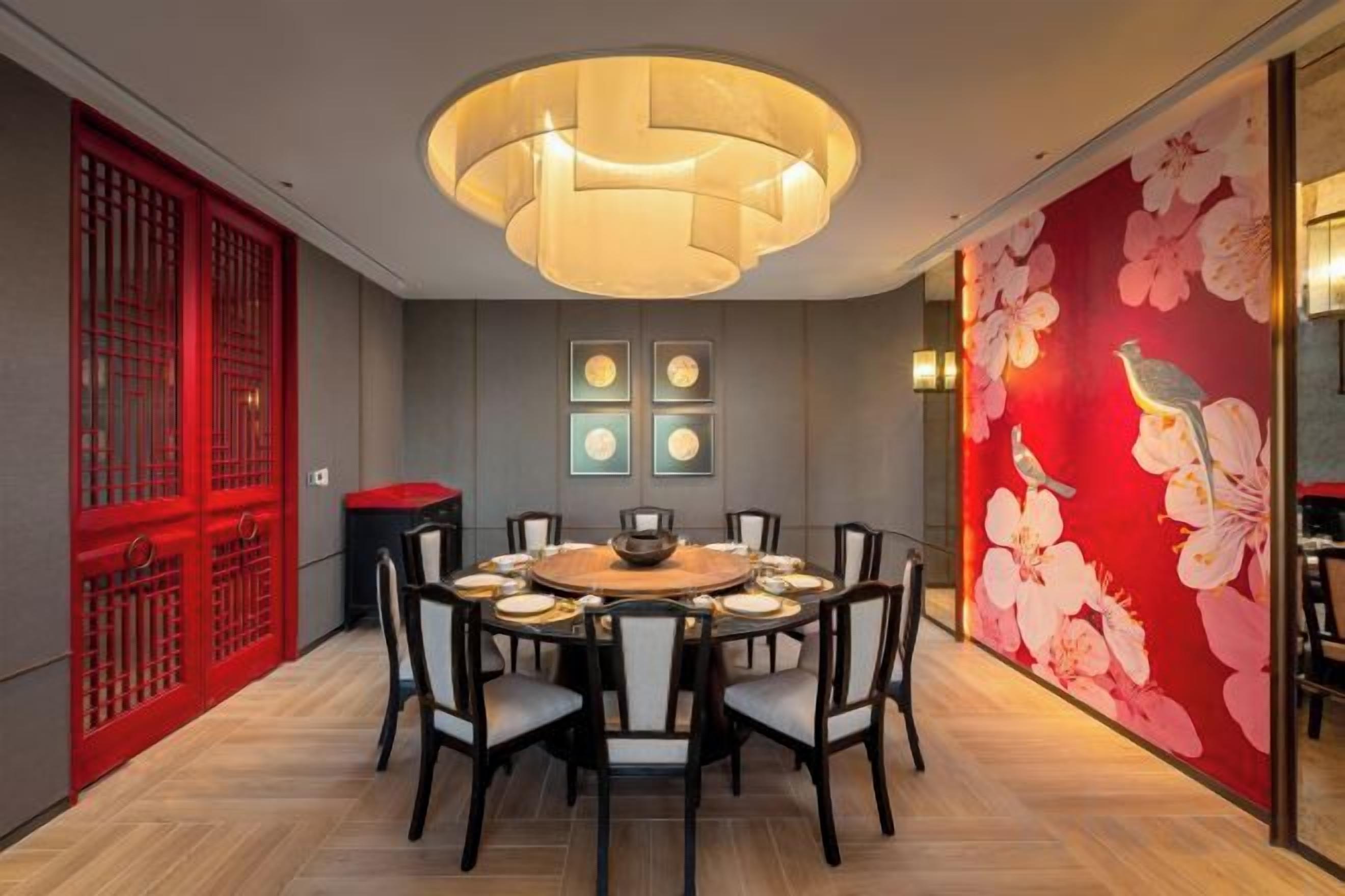 A dining area with a round table, chairs, a central light fixture, and a red decorative wall with floral patterns.
